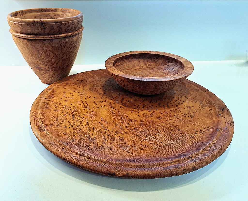 A set of wooden bowls and pots made from burl wood, featuring a natural finish and intricate grain patterns. The bowls are on a flat wooden plate, showcasing the unique texture and color of the wood.