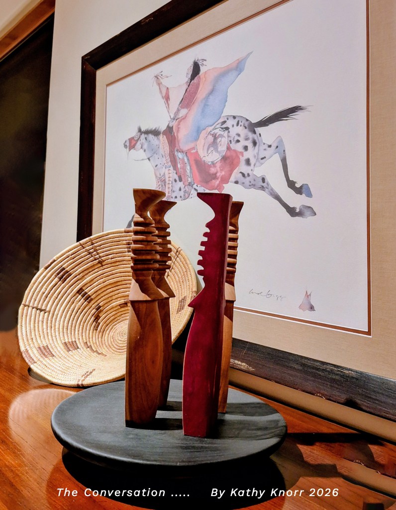A wooden sculpture titled 'The Conversation' by Kathy Knorr, featuring two abstract figures in contrasting colors, placed on a circular black base. In the background, there is a framed watercolor painting of a rider on a horse, accompanied by a woven decorative plate.